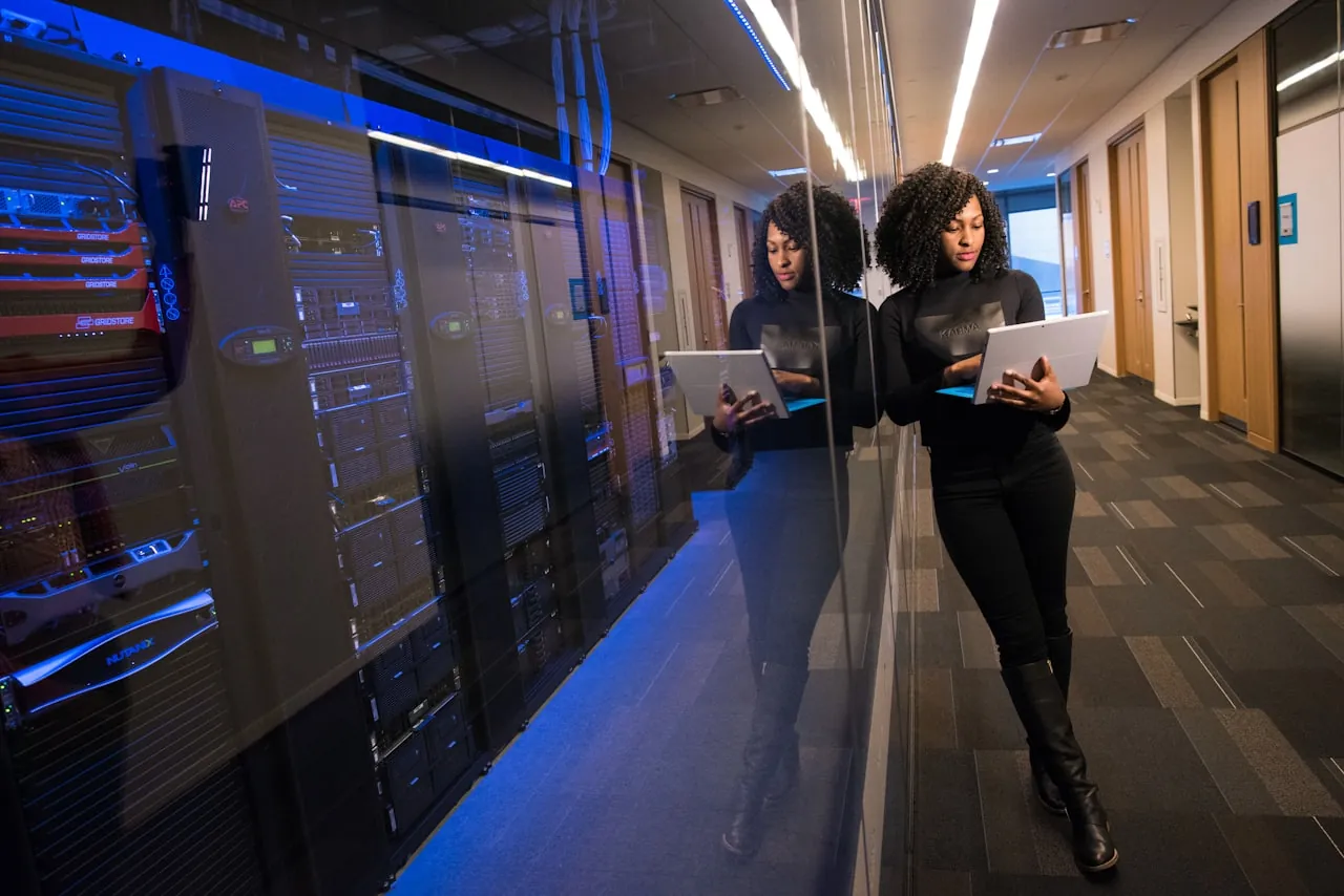 An engineer with a laptop in a data center corridor, beside server racks behind glass